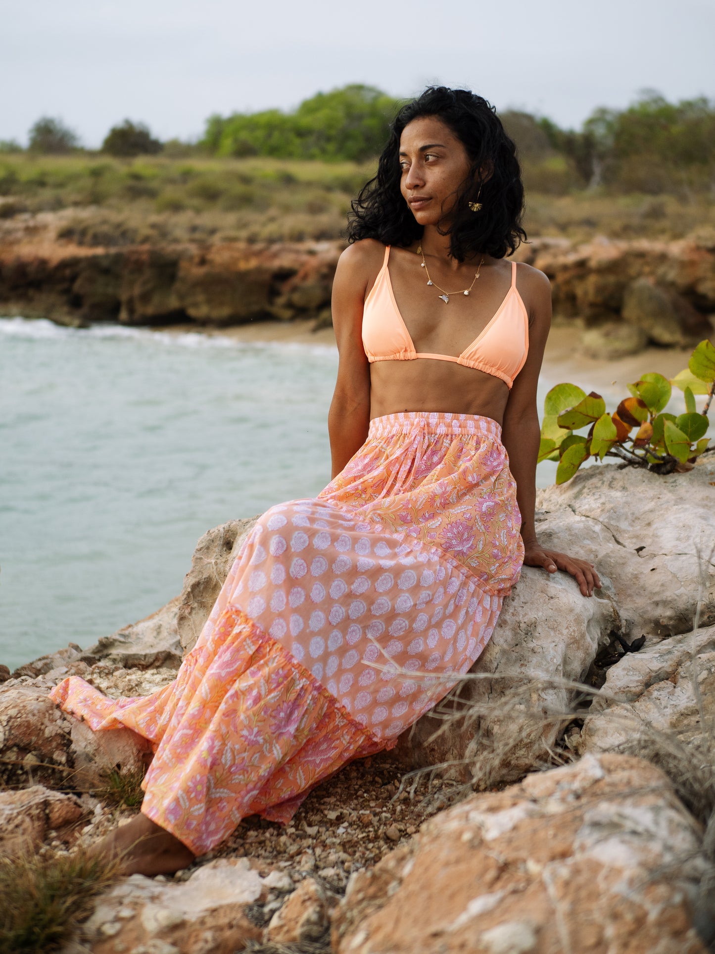 Woman in a peach bikini top and block printed skirt sitting on rocks by a body of water.